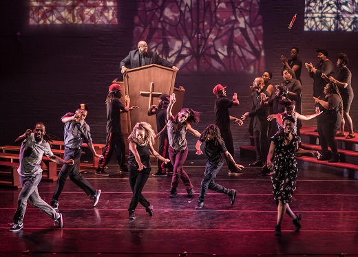 Onstage, a group of smiling Puremovement dancers in a cross step in the foreground; Robinson Jr. on a raised pulpit in the background; and Alonzo Chadwick & Friends clapping and singing on choir risers.